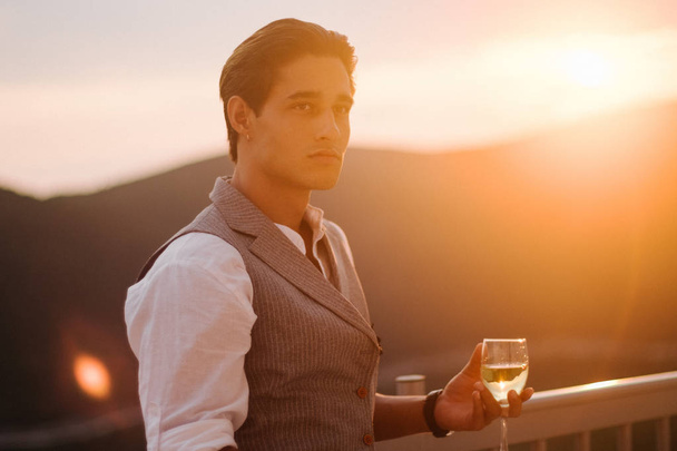 Attractive young fashion man in a suit supper on the terrace and drinks white wine next to the villa. - Foto, imagen