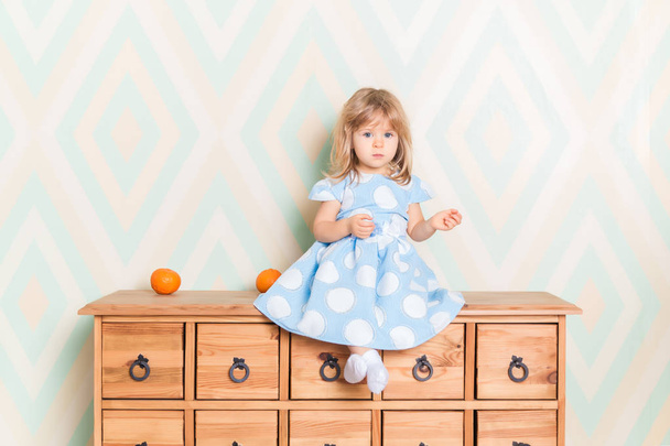 A little baby girl in her room sitting cross-legged on chest of drawers with tangerines on the rhomb wallpaper background. Child in blue polka dot dress and white socks attentively looking at camera - Photo, image