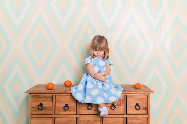 A little baby girl in her room sitting cross-legged on chest of drawers with tangerines on the rhomb wallpaper background. Child in blue polka dot dress and white socks attentively looking down - Foto, immagini