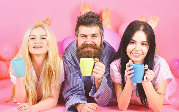 Threesome relax in morning with coffee. Lovers concept. Man and women, friends on smiling faces lay, pink background. Lovers in love drink coffee in bed. Man and women in domestic clothes, pajamas - Foto, immagini