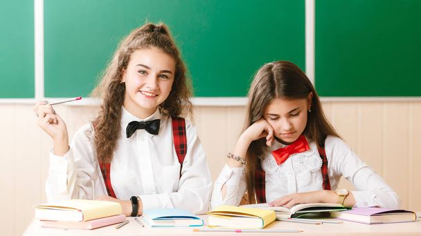 две школьницы-брюнетки сидят в классе за партой с книгами на фоне черной доски.
 - Фото, изображение