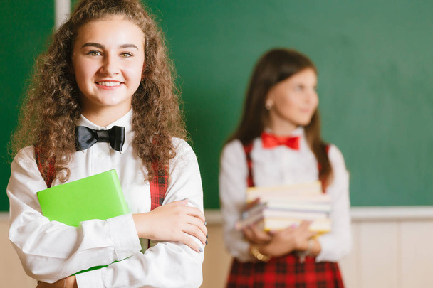 two brunette schoolgirls in school red uniforms stand in a classroom with books.schoolgirl looking at the camera and smiling - Photo, Image