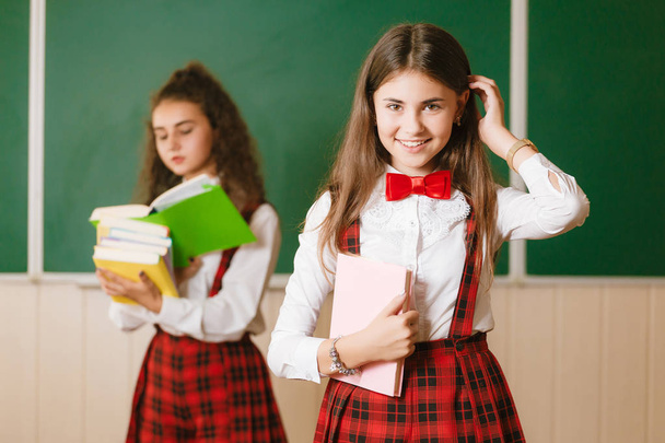 dos colegialas morenas en uniforme rojo de la escuela están en un aula con books.schoolgirl mirando a la cámara y sonriendo
 - Foto, Imagen