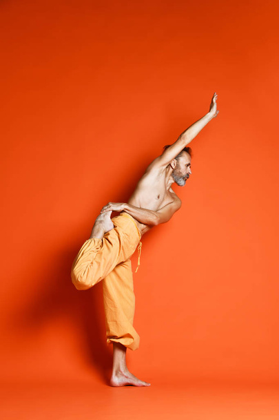 Old man practicing yoga doing stretching exercises against orange background - Фото, изображение