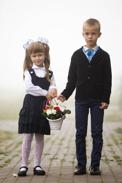 Full-length portrait of two children, serious sad boy in embroidered shirt and smiling first grader girl in school uniform and white bows holding flower composition on blurred copy space background. - Zdjęcie, obraz