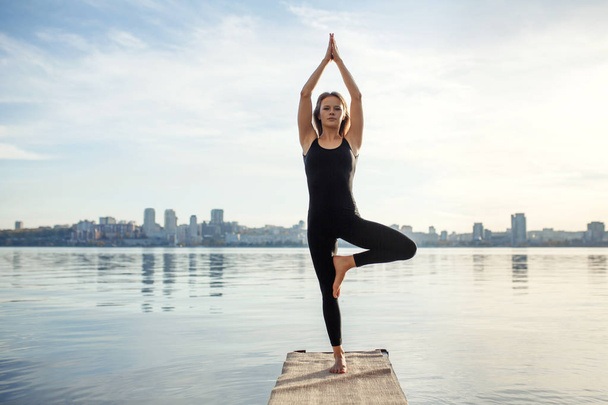 Young woman practicing yoga exercise at quiet wooden pier with city background. Sport and recreation in city rush. - Фото, зображення