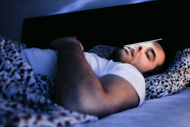 Νεαρός άνδρας χρησιμοποιώντας το smartphone στο κρεβάτι τη νύχτα - Φωτογραφία, εικόνα