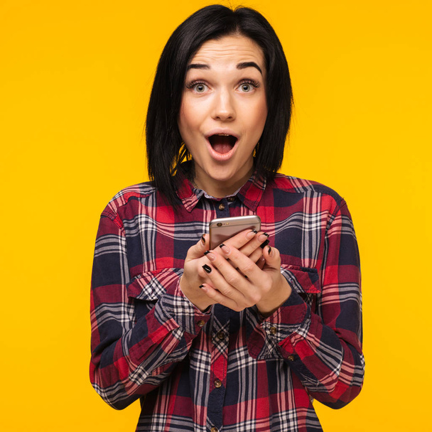 Excited laughing woman in plaid shirt standing and using mobile phone over yellow background - Image - Photo, Image