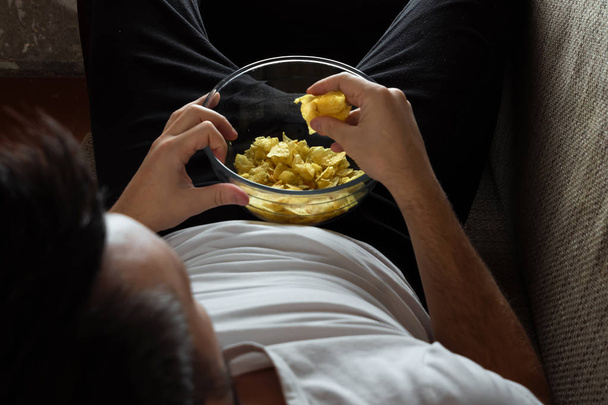 The guy in the shirt is lying on the couch, eating chips and watching a sports channel. The concept of laziness, frustration, procrastination, the person at home. - Φωτογραφία, εικόνα