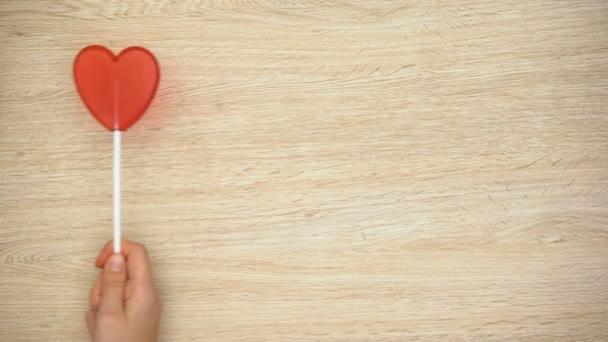 Hand holding heart-shaped lollipop against wooden background, love, top view - Felvétel, videó