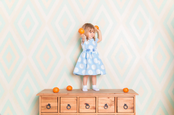 Toddler child girl in blue dress standing on wooden chest of drawers and holding fresh orange mandarins in her hands and going to throw them on rhomb wallpaper background. Winter holidays or christmas - Foto, afbeelding