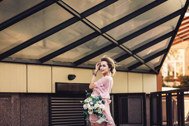 ελκυστική νεαρή κοπέλα σε ένα κοντό φόρεμα με ένα μπουκέτο λουλούδια που ποζάρουν στο δρόμο κοντά στον τοίχο του κτιρίου. - Φωτογραφία, εικόνα
