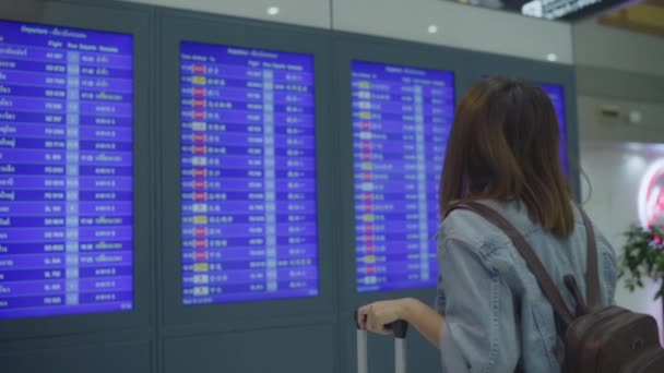 スローモーション - 国際空港の出発ゲート ターミナル ホールで荷物と彼女のフライトをチェック情報板を見て幸せなアジア女。空港のコンセプトで幸せなライフ スタイルの女性. - 映像、動画