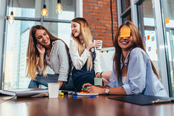 Two girls laughing at their friend with a sticky note on her face. Group of female students relaxing having fun in classroom during a break - Foto, imagen