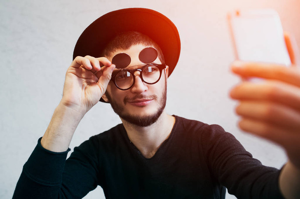 Portrait of young man taking selfie, standing over white background. Dressed in black, wearing sunglasses and hat - Foto, imagen