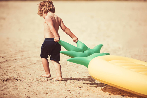 Small boy child pull air mattress on sand. Summer vacation and travel to ocean. Pineapple inflatable mattress, activity and joy. Maldives or Miami beach. little kid on Caribbean sea in Bahamas. - 写真・画像