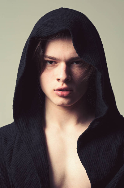 Guy on confident face in black hood, grey background. Man with smooth skin on face, neck and chest looking at camera. Masculinity concept. Macho in black bathrobe with hood, close up - Φωτογραφία, εικόνα