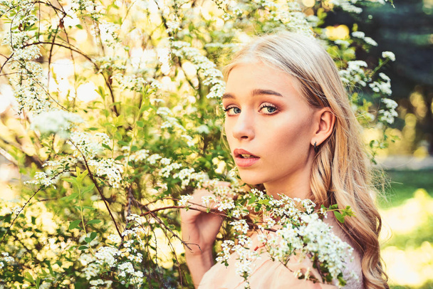 Girl on dreamy face, tender blonde near branches with white flowers, nature background. Lady walks in park on sunny spring day. Spring bloom concept. Young woman enjoy flowers in garden. - Fotó, kép
