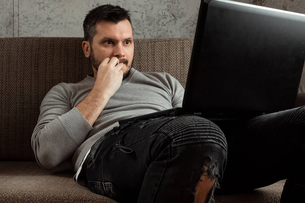 A man watches an adult video on a laptop while sitting on the couch. The concept of porn, men's needs, pervert, lust, desire, loneliness. - Fotó, kép