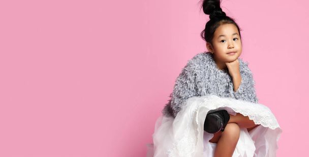 Young asian girl kid in long white skirt, grey fluffy sweater and black shoes sitting legs crossed and looks direct. Lean her chin on her fist. - Zdjęcie, obraz