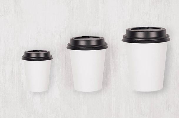 Coffee mockup - set of three different size white paper cups and blank black cap on white wood board, top view. Modern elegant concept for branding identity, advertising, design. - Foto, Imagen