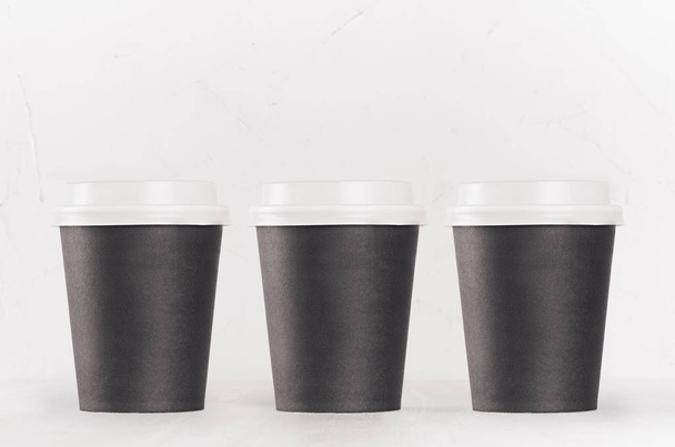 Coffee mockup - set of three blank black paper cups with white cap on white wood table, coffee shop interior. Modern elegant concept for branding identity, advertising, design. - Foto, afbeelding
