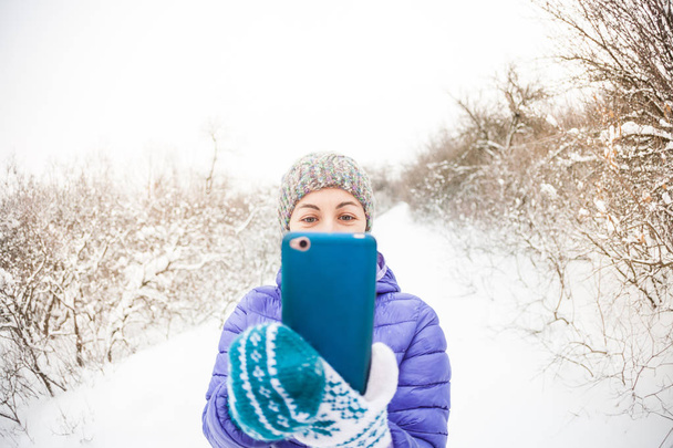 Smartphone. Μια γυναίκα κάνει μια φωτογραφία σε ένα κινητό τηλέφωνο μέσα στο δάσος του χειμώνα. Ένα χαμογελαστό κορίτσι σε ένα πλεκτό καπέλο κάνει selfie φόντο ένα πάρκο χιόνι που καλύπτει. - Φωτογραφία, εικόνα