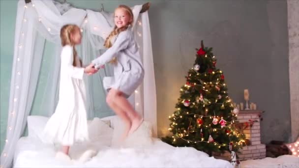 Vista en cámara lenta de dos chicas descalzas sonrientes con trenzas y en ropa de dormir cogidas de la mano, saltando sobre la cama. Chimenea y árbol de Navidad están en el fondo - Metraje, vídeo