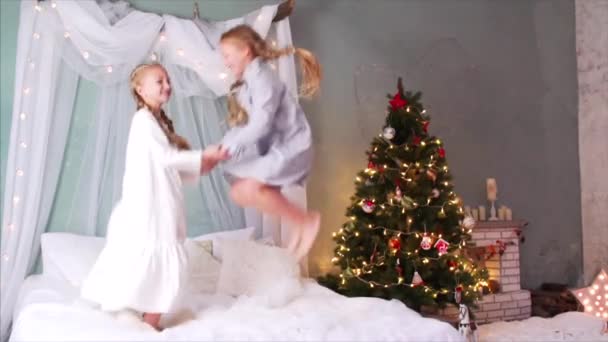 Άποψη αργής κίνησης δύο χαμογελαστών γυμνόστηθων κοριτσιών με κοτσίδες και νυχτικά που κρατιούνται χέρι χέρι, πηδώντας ψηλά στο κρεβάτι. Τζάκι και χριστουγεννιάτικο δέντρο είναι στο παρασκήνιο - Πλάνα, βίντεο