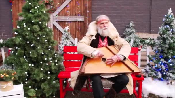 Bianco barbuto vecchio in costume nazionale russo è seduto sulla panchina vicino agli alberi di Natale, giocare dulcimer e cantare - Filmati, video