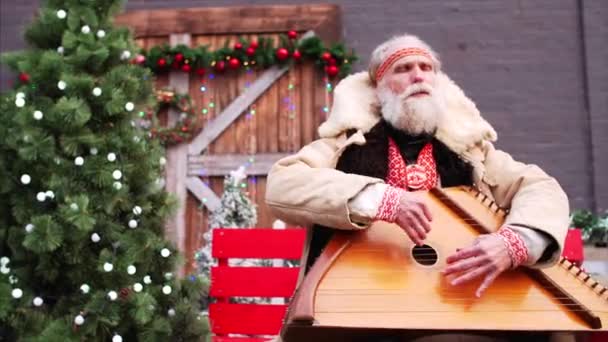 Retrato deslizante de anciano con barba blanca en traje ruso nacional rústico sentado en el banco cerca de árboles de Navidad, jugando dulcimer y cantando muy emocionalmente - Metraje, vídeo