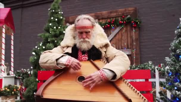 Retrato deslizante de anciano con barba blanca en traje ruso nacional rústico entre las decoraciones de Navidad jugando dulcimer - Metraje, vídeo