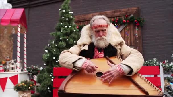 Retrato deslizante de anciano con barba blanca en traje ruso nacional rústico entre las decoraciones de Navidad jugando dulcimer y cantando emocionalmente - Metraje, vídeo
