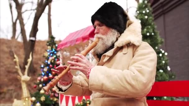 Portrét bílého vousatého starce v teplém kabátě a černém klobouku hrající dřevěnou dýmku na lavičce mezi vánočními ozdobami a vánočními stromky - Záběry, video