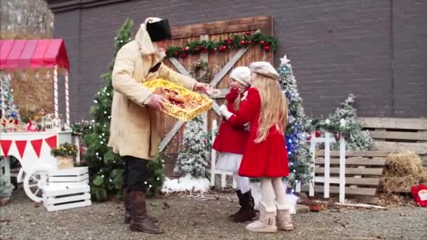 Bílý vousatý stařec v kabátě a klobouku dává domácí krekry pro dvě dívky v červených kabátech na vánočním veletrhu - Záběry, video