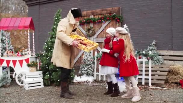 Hombre viejo barbudo blanco en abrigo y sombrero está vendiendo galletas caseras para dos niñas en abrigos rojos en la feria de Navidad - Imágenes, Vídeo