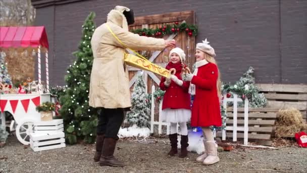 Bílý vousatý stařec v kabátě a klobouku dává domácí krekry pro dvě dívky v červených kabátech na vánočním veletrhu - Záběry, video