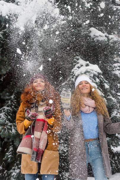 Δύο Καυκάσιος αδερφές κορίτσια με σγουρά μαλλιά στέκεται ανάμεσα στα δέντρα καλυμμένα με χιόνι του χειμώνα και ρίχνοντας επάνω χιόνι. Χειμώνα απόλαυση και διασκέδαση - Φωτογραφία, εικόνα