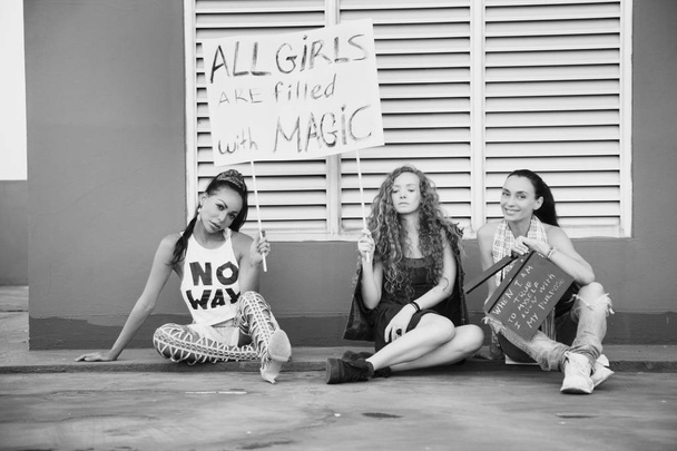 Die Mädchen sitzen mit einem Plakat auf der Straße. Weltfrauentag. Frauen unterschiedlicher Nationalitäten, Kulturen und Geschlechtsidentität. Kampf um Freiheit, Unabhängigkeit, Gleichheit - Foto, Bild
