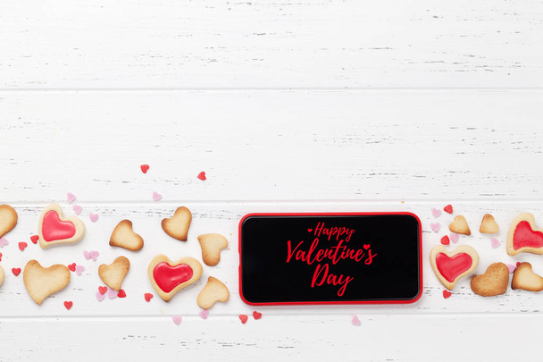 Открытка на день Святого Валентина с печеньем в форме сердца и смартфоном на деревянном фоне. Вид сверху с местом для приветствия или приложения для смартфона. Плоский лежал
 - Фото, изображение