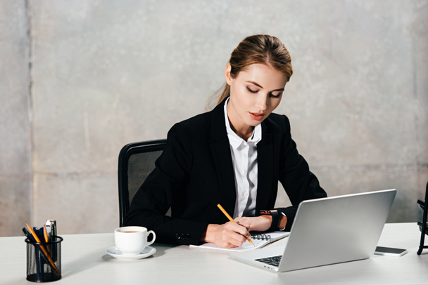 young concentrated businesswoman sitting at workplace and making notes - Photo, Image