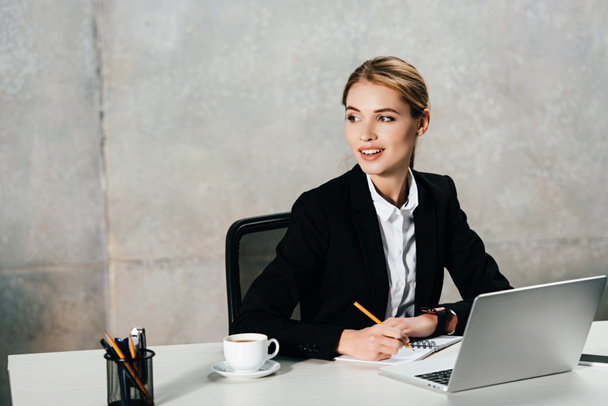 smiling businesswoman sitting at workplace and writing in notebook - Фото, изображение