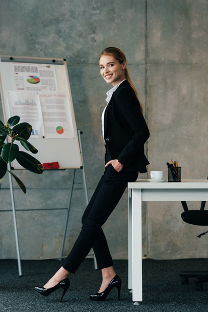 smiling businesswoman standing by work desk and keeping hands in pockets - Fotografie, Obrázek