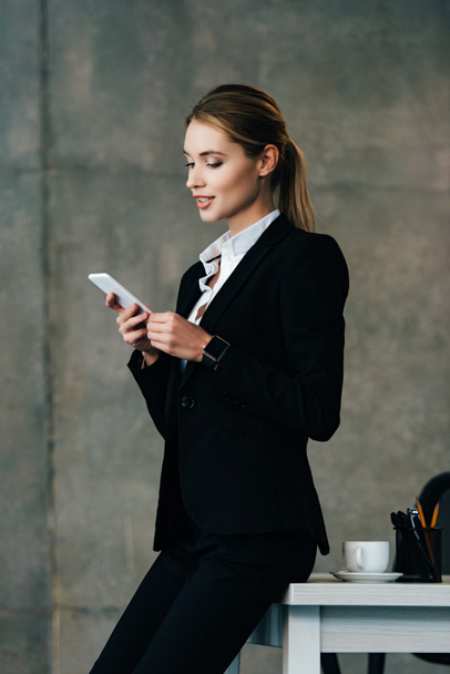 smiling businesswoman standing by desk and using smartphone in office - Foto, Imagen