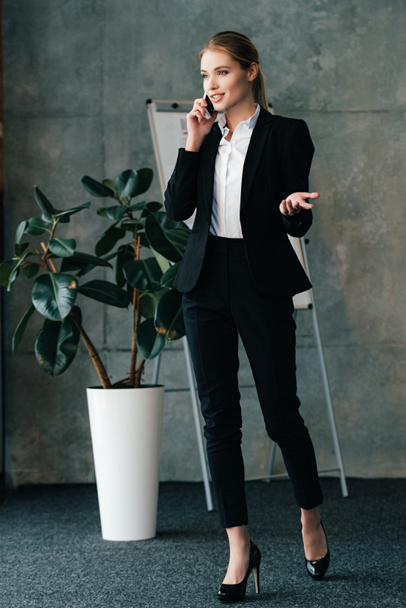 attractive smiling businesswoman talking by phone while standing near large flower pot - Foto, Imagem