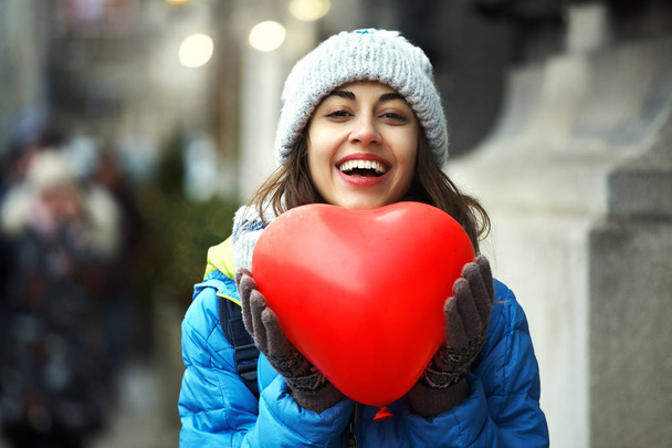 happy young woman in warm winter clothes, knitted cap and scarf is standing outdoors with a heart shaped red balloons - Photo, Image