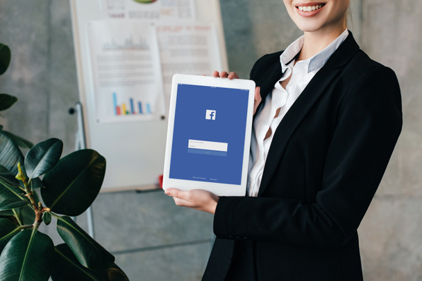 partial view of pretty smiling businesswoman holding digital tablet with facebook on screen - Zdjęcie, obraz