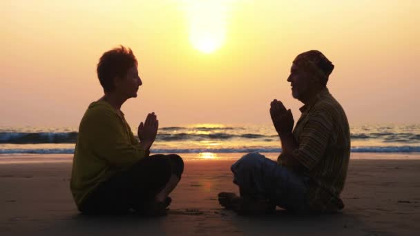 Silhouet van senior paar zit en samen mediteren op zandstrand. Oudere man en vrouw zit aangezicht tot aangezicht in Turkse pose met gesloten ogen. - Video