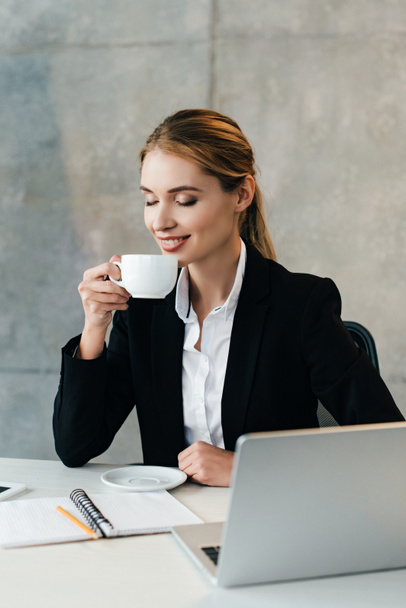 smiling businesswoman enjoys drinking coffee with closed eyes while sitting at workplace in office - Fotografie, Obrázek