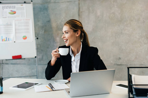 Beautiful laughing businesswoman sitting at workplace and drinking coffee  - Photo, Image